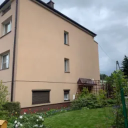 Widok z poziomu ogrodu na trzypiętrowy budynek z beżową elewacją, brązowymi oknami i dachem, oraz zadbanym trawnikiem i rabatami kwiatowymi w odcieniach czerwieni, żółci, fioletu i bieli.