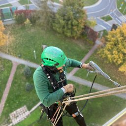 4Invest - Pracownik w zielonym kasku, zabezpieczony linami, używa myjki ciśnieniowej na wysokości, widok z góry na budynek i otoczenie z rondem.