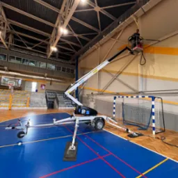 Pracownik na podnośniku koszowym w hali sportowej, wykonujący prace na wysokości przy suficie, widoczny parkiet, trybuny i bramka do piłki ręcznej.