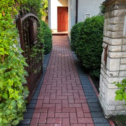 4Invest - Czerwona kostka brukowa prowadząca do wejścia domu, otoczona zielenią i elementami architektury ogrodowej.