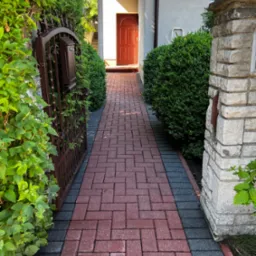 Czerwona kostka brukowa prowadząca do wejścia domu, otoczona zielenią i elementami architektury ogrodowej.