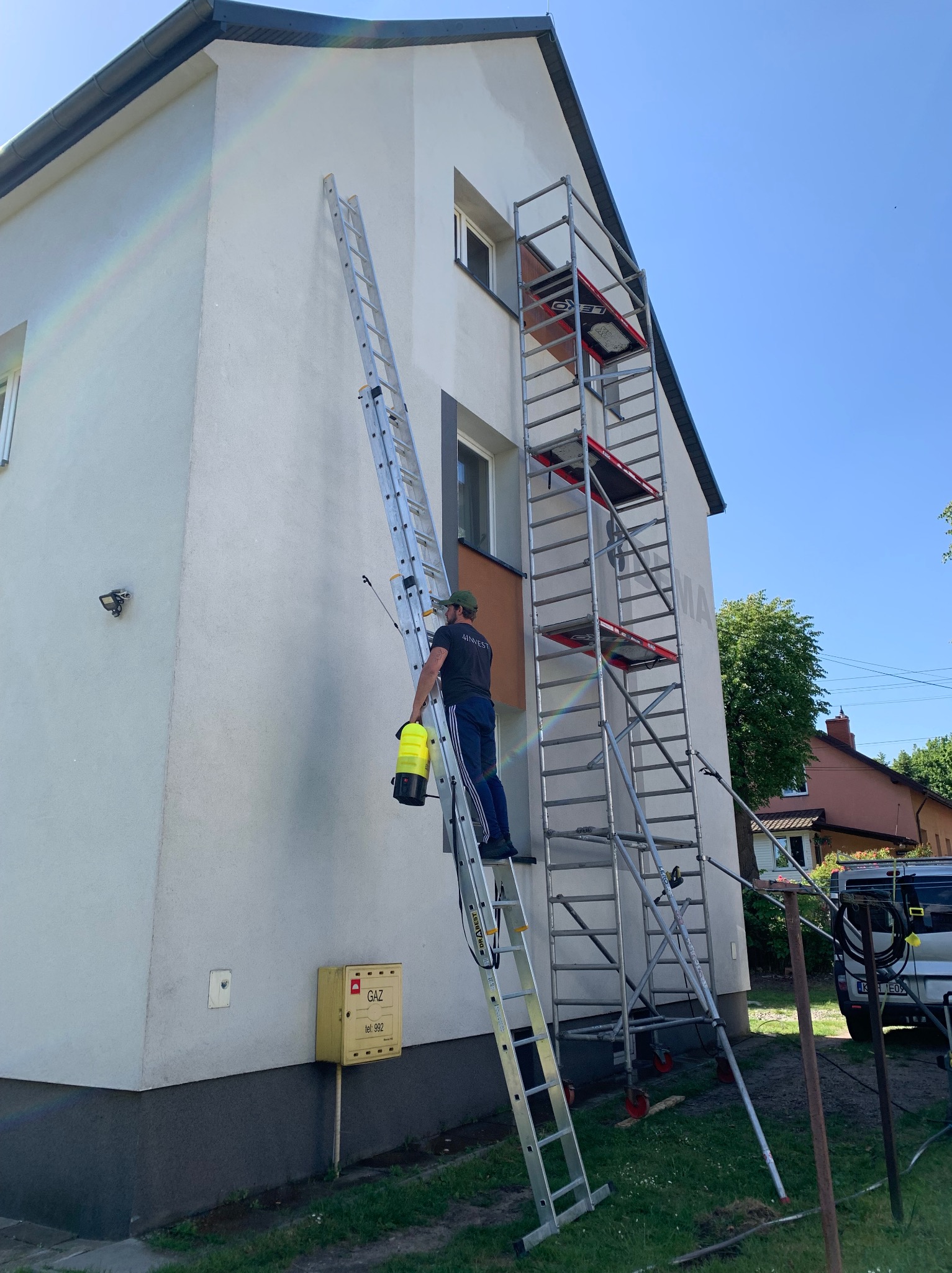 Pracownik na drabinie z opryskiwaczem, przygotowujący się do mycia elewacji dwupiętrowego domu z rusztowaniem, widoczna skrzynka gazowa.