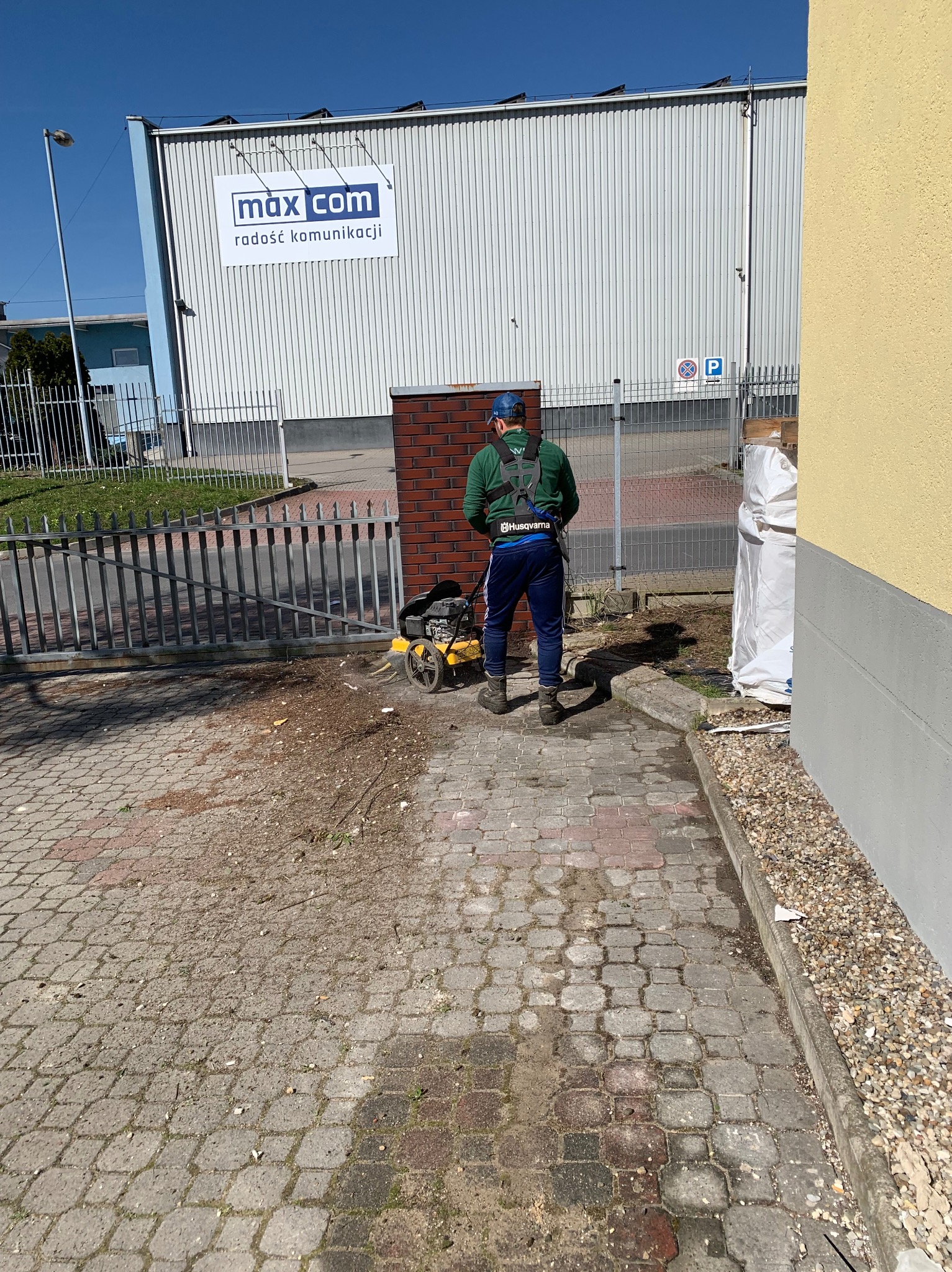 Mężczyzna w zielonej bluzie i niebieskich spodniach używa żółtej krawędziarki do trawy na kostce brukowej przed budynkiem z logo 'max com', widoczny znak zakazu parkowania.