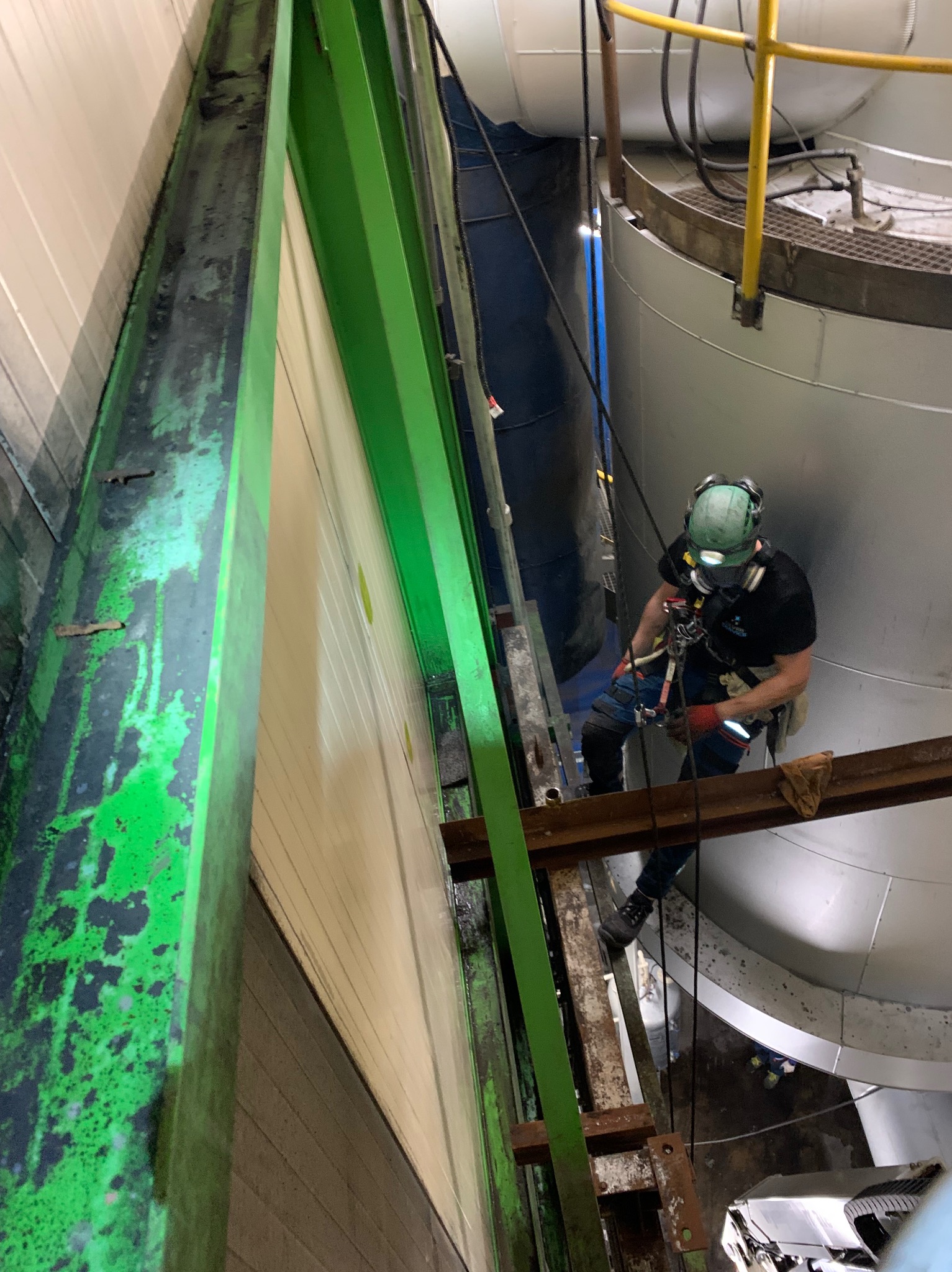 Pracownik w uprzęży, kasku i masce, na wysokości, na tle przemysłowych zbiorników i konstrukcji stalowych, prawdopodobnie podczas inspekcji lub czyszczenia.