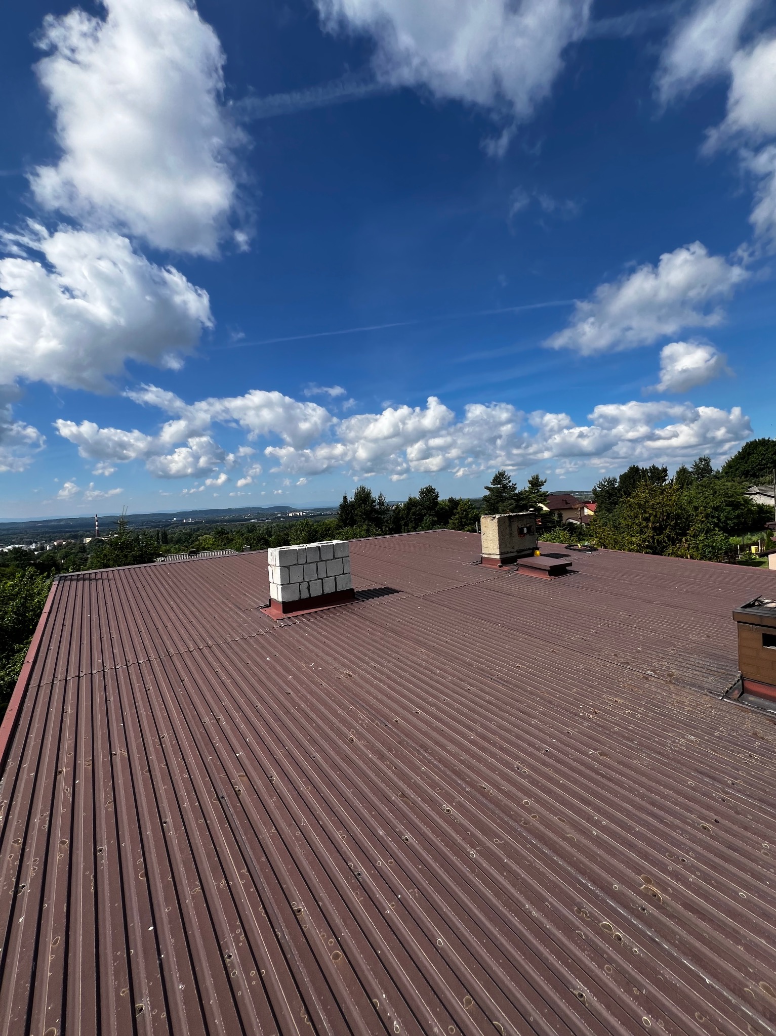 Czerwony dach z blachy trapezowej z widocznymi kominami, na tle błękitnego nieba z białymi chmurami i zalesionego terenu.