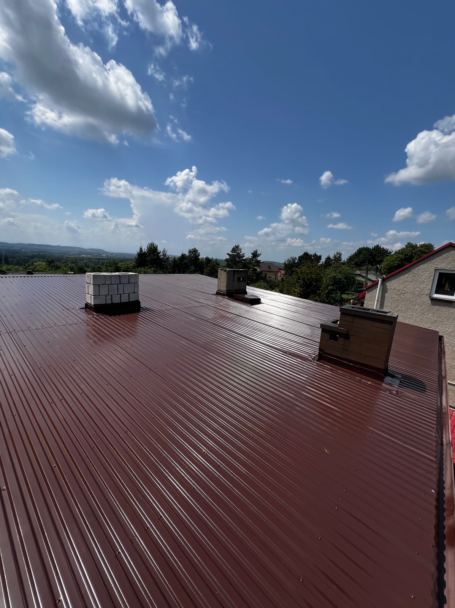 Czerwony dach z blachy trapezowej, trzy kominy, widok na okolicę w słoneczny dzień.