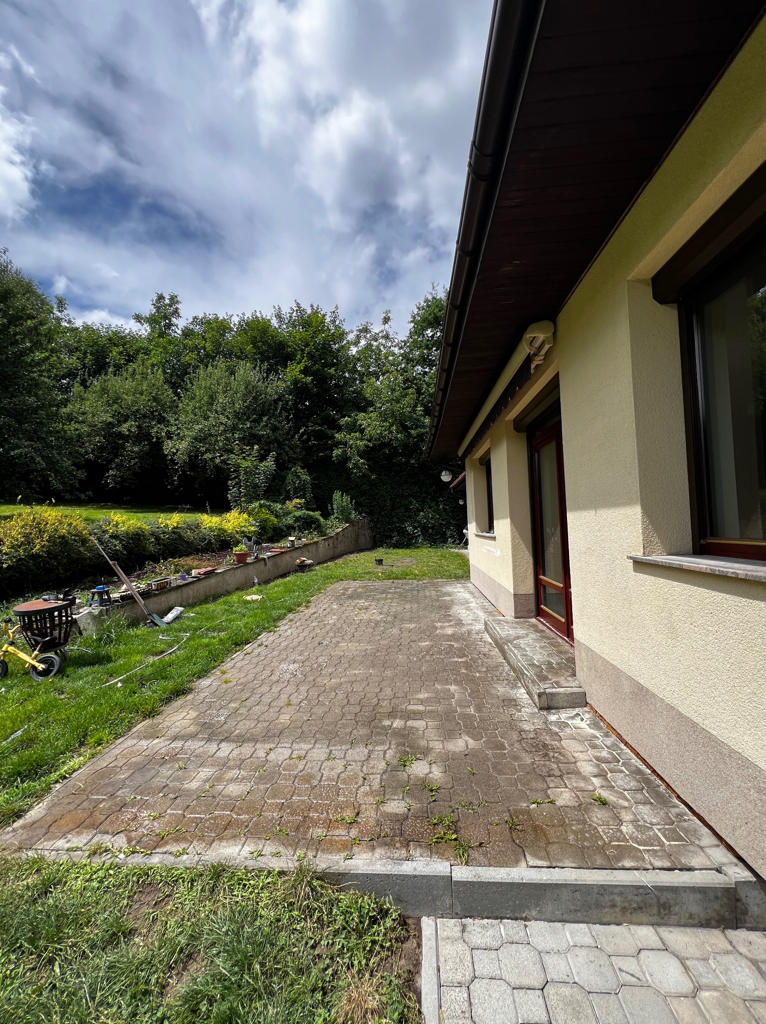 Częściowo wyczyszczony, mokry chodnik z kostki brukowej przy elewacji domu, z widocznym odrostem chwastów w spoinach, ogród z zielenią w tle i żółty rowerek dziecięcy po lewej.