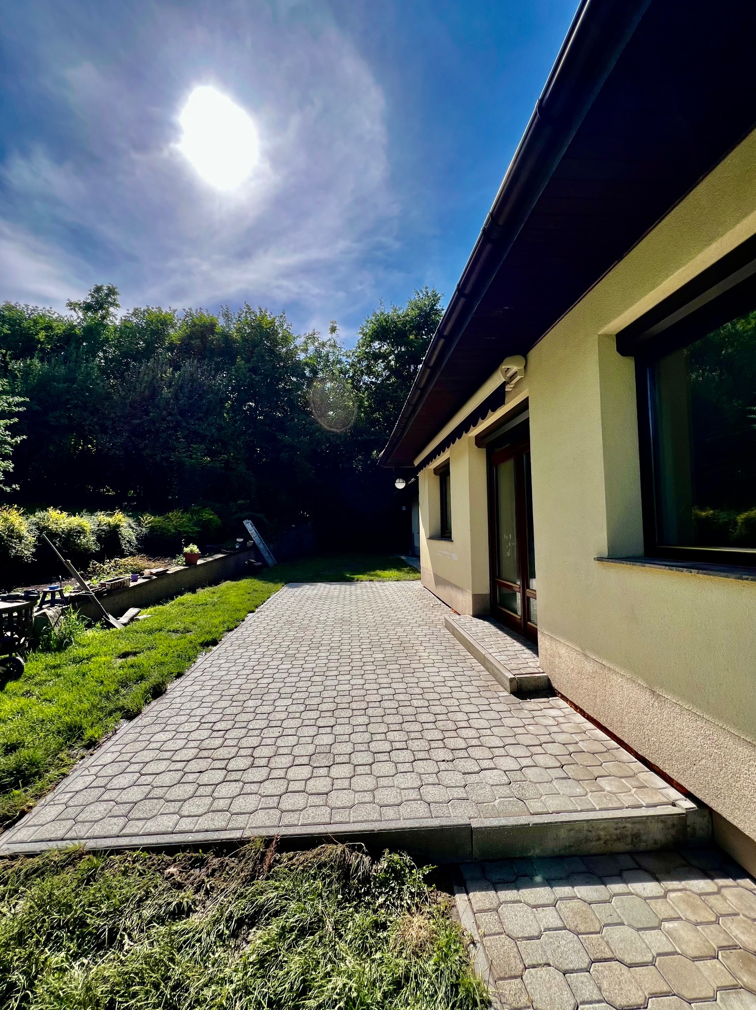 Nowo ułożona, szara kostka brukowa tworzy patio przylegające do jasnego domu z ciemnymi oknami i dachem, widok na zielony trawnik i drzewa w tle, jasne słońce na niebie.