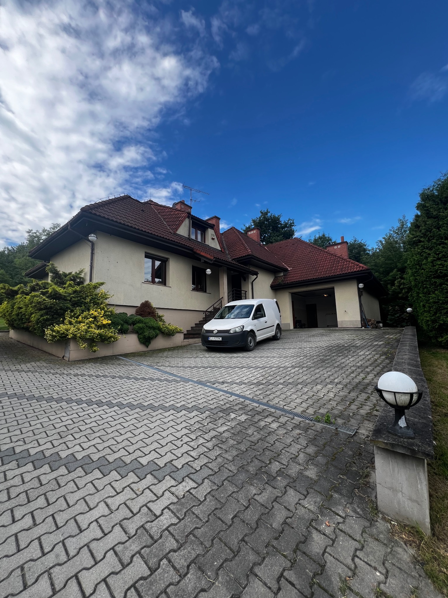 Jasny dom jednorodzinny z czerwoną dachówką, podjazd z kostki brukowej i biały samochód dostawczy zaparkowany przed garażem w słoneczny dzień.