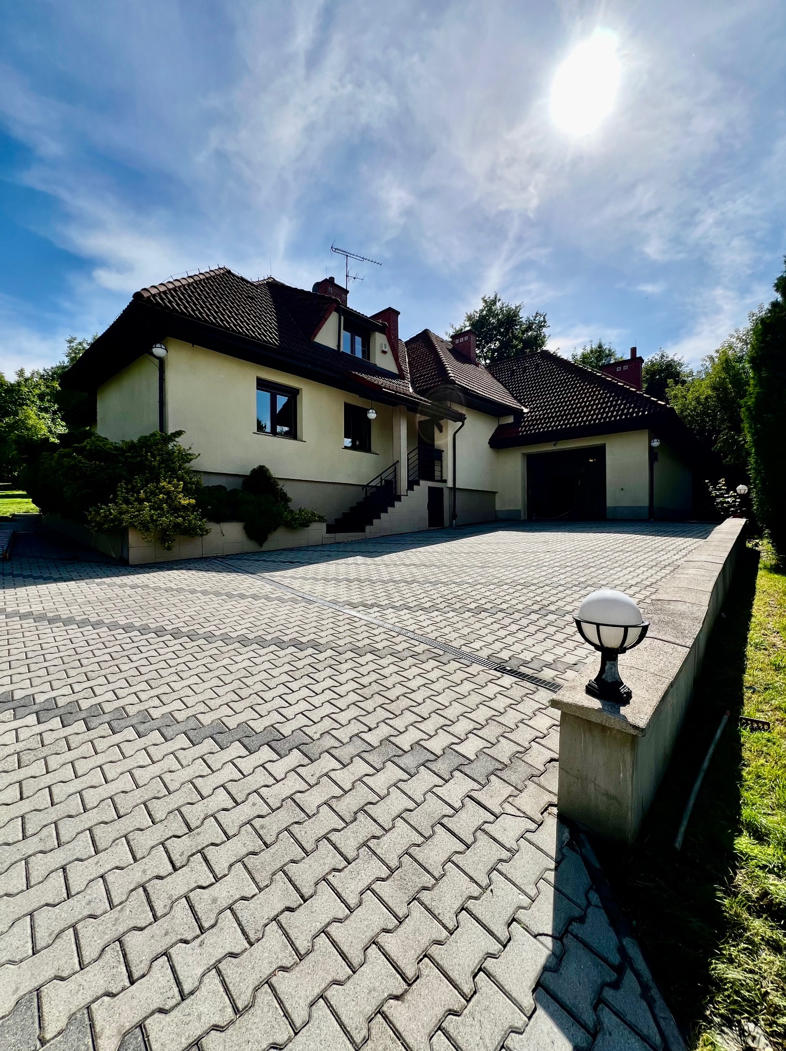 Dwupiętrowy dom jednorodzinny z garażem, widoczny z perspektywy podjazdu wyłożonego szarą kostką brukową, oświetlonego kulistą lampą ogrodową.