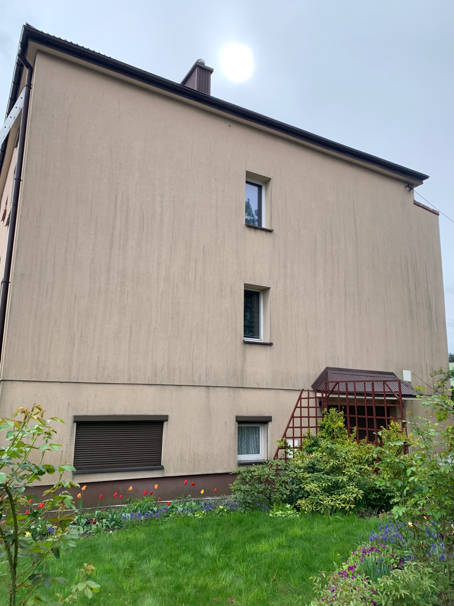 Trzypiętrowy budynek z beżową elewacją, widoczne zabrudzenia i zacieki, brązowe rynny i okna, ogródek z kwiatami i krzewami u podnóża ściany, pochmurne niebo.