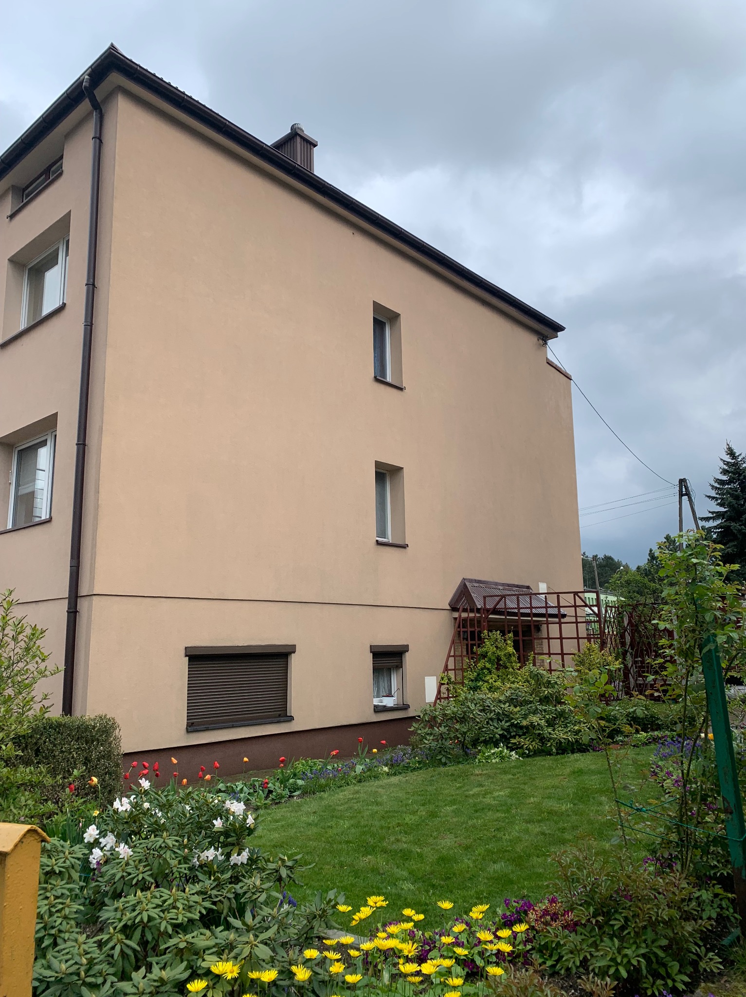 Widok z poziomu ogrodu na trzypiętrowy budynek z beżową elewacją, brązowymi oknami i dachem, oraz zadbanym trawnikiem i rabatami kwiatowymi w odcieniach czerwieni, żółci, fioletu i bieli.