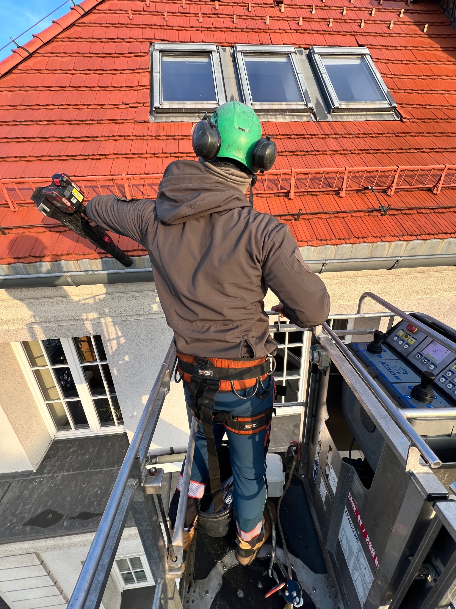 Pracownik w zielonym kasku i słuchawkach na platformie roboczej, z urządzeniem w ręku, na tle czerwonego dachu z oknami dachowymi.