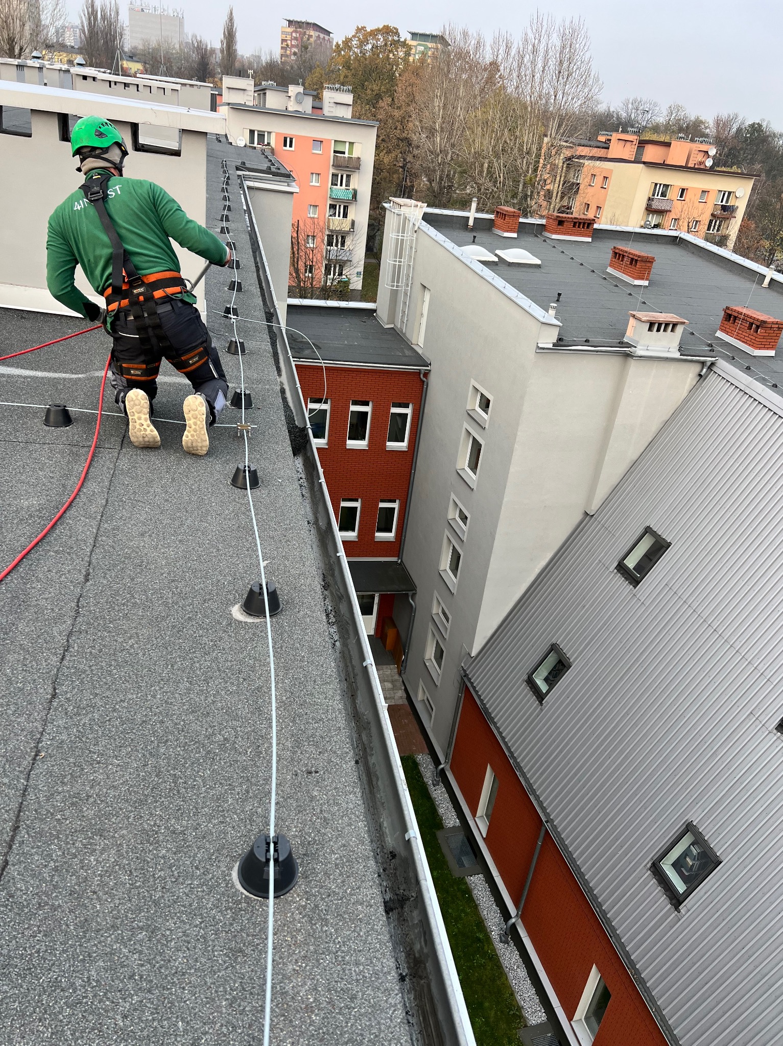 Pracownik w zielonym kombinezonie i kasku, zabezpieczony linami, wykonuje prace na płaskim dachu budynku z widocznymi elementami zabezpieczającymi przed upadkiem, w tle zabudowa miejska.