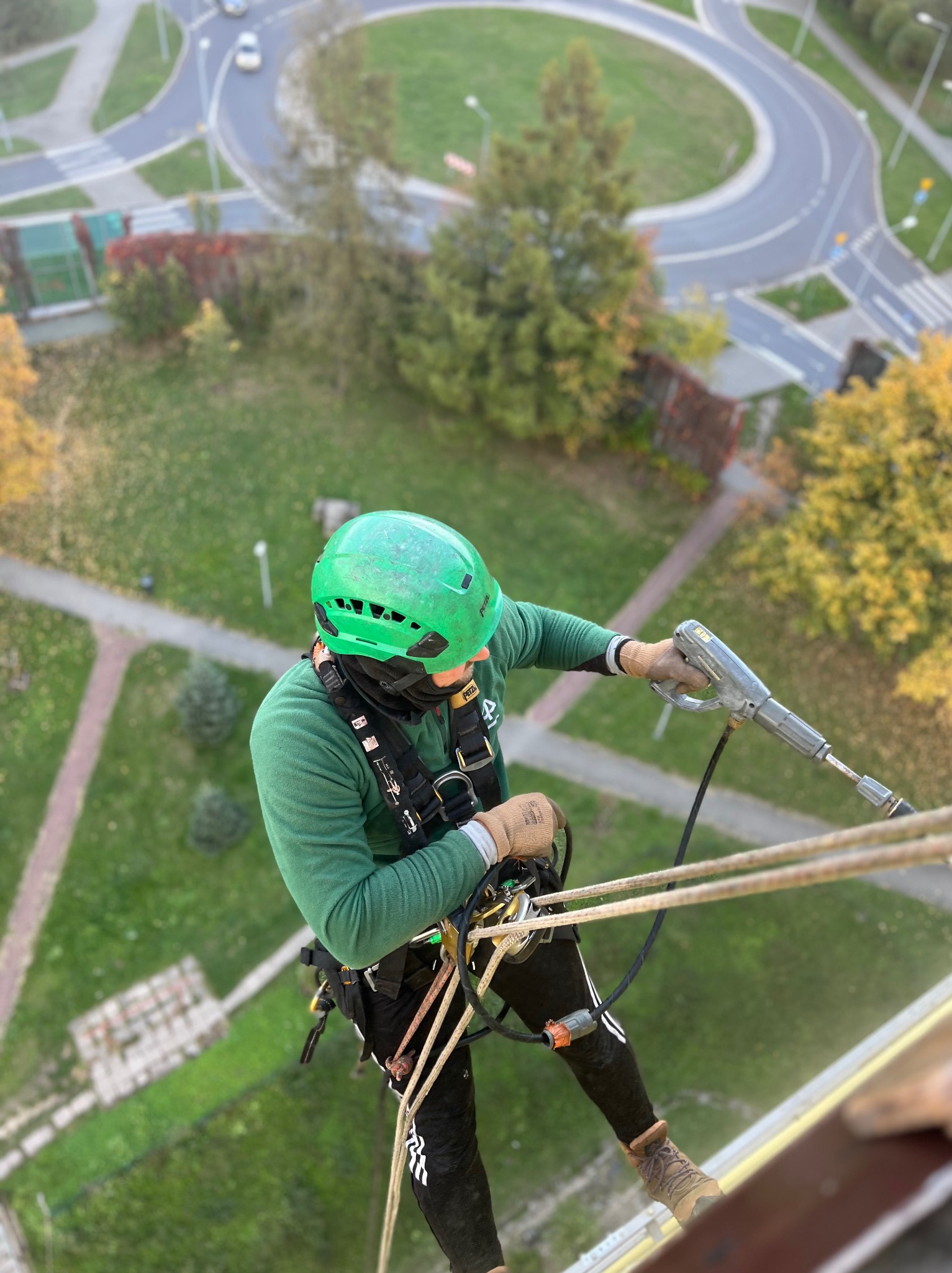 Pracownik w zielonym kasku, zabezpieczony linami, używa myjki ciśnieniowej na wysokości, widok z góry na budynek i otoczenie z rondem.
