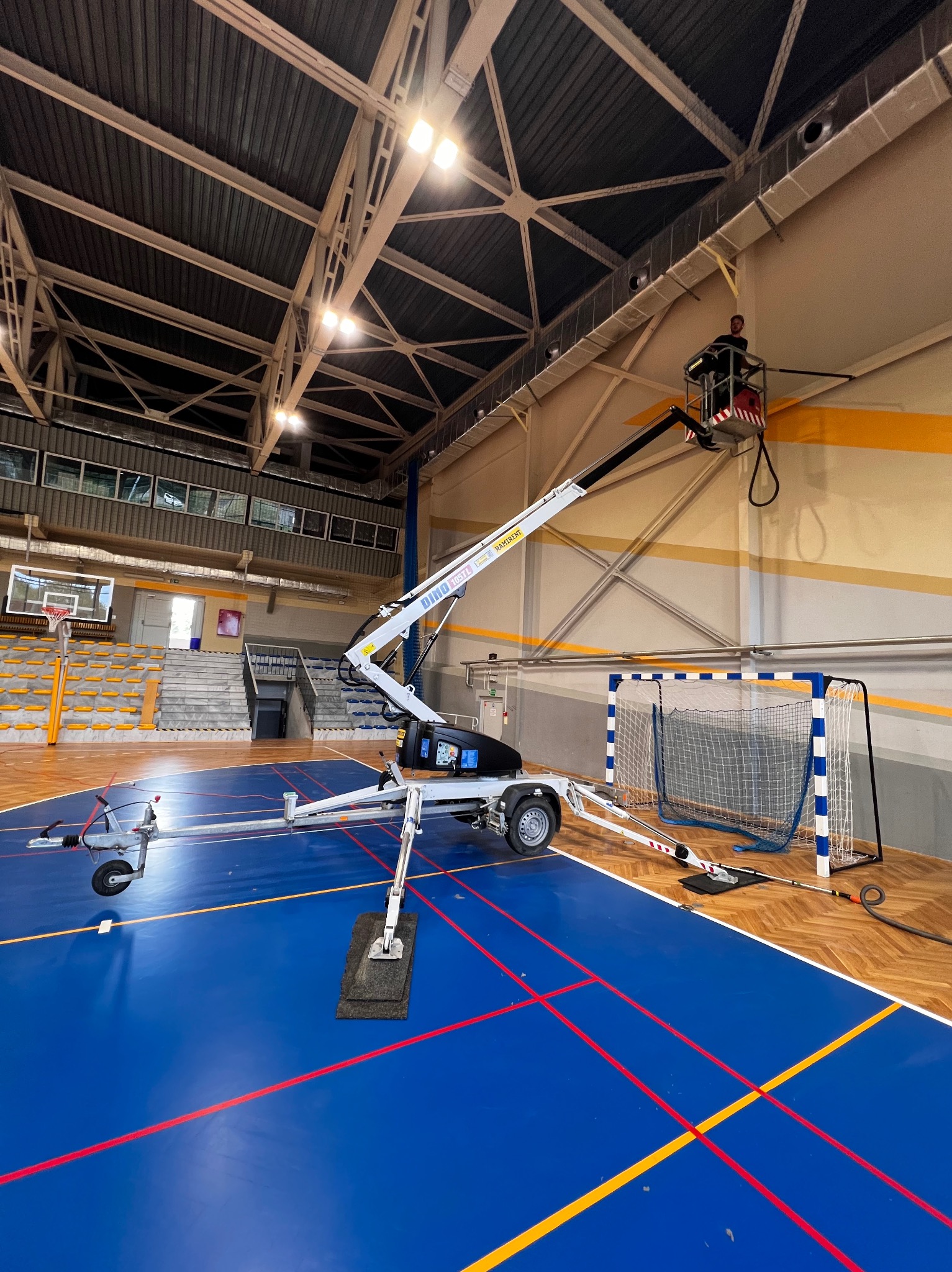 Pracownik na podnośniku koszowym w hali sportowej, wykonujący prace na wysokości przy suficie, widoczny parkiet, trybuny i bramka do piłki ręcznej.