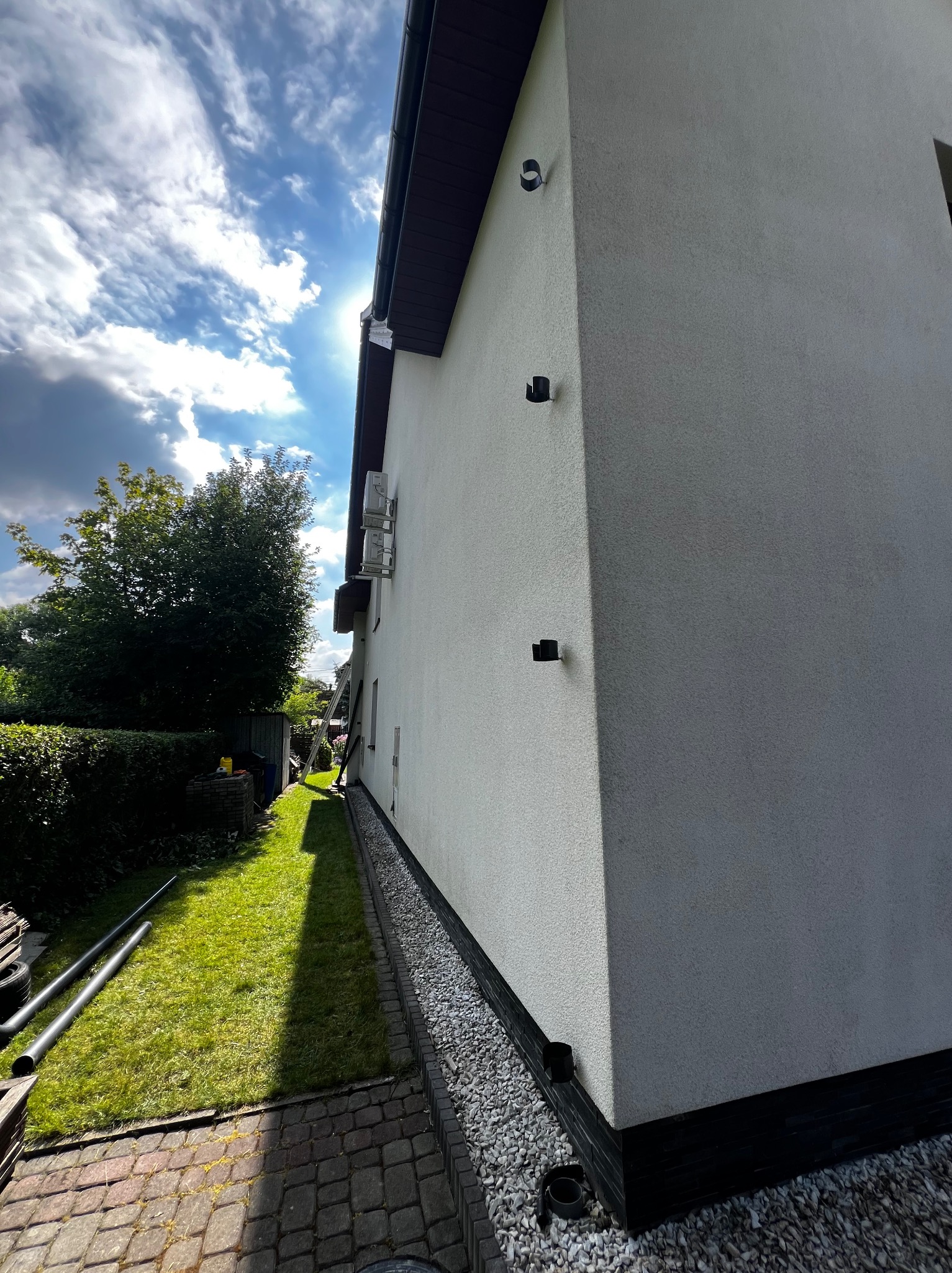 Boczny widok domu z białą elewacją w słoneczny dzień, widoczna zieleń trawnika, rury spustowe i drabina oparta o ścianę.