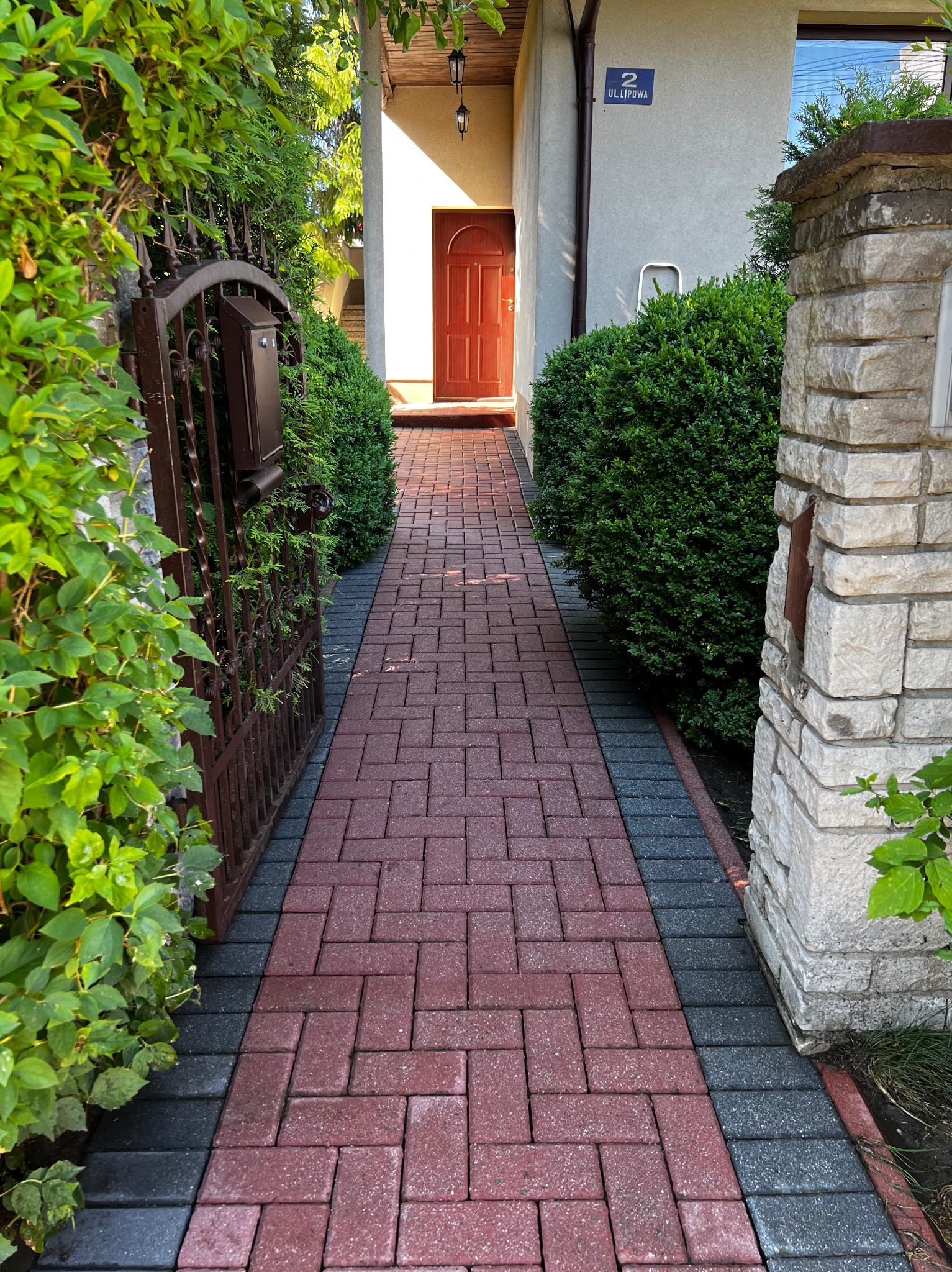 Czerwona kostka brukowa prowadząca do wejścia domu, otoczona zielenią i elementami architektury ogrodowej.