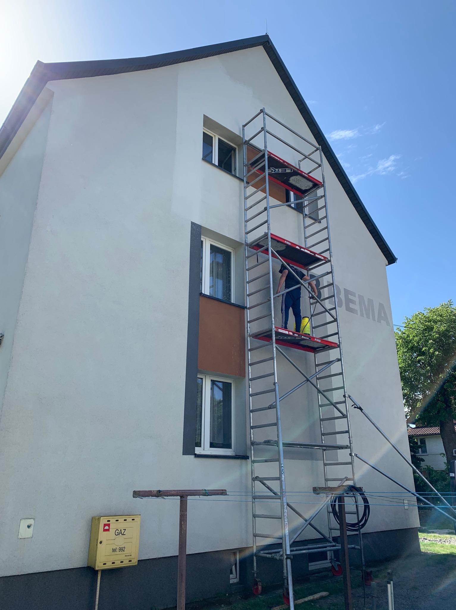 Pracownik na rusztowaniu czyści elewację budynku z napisem, używając myjki ciśnieniowej; widoczna skrzynka gazowa i fragmenty zieleni.