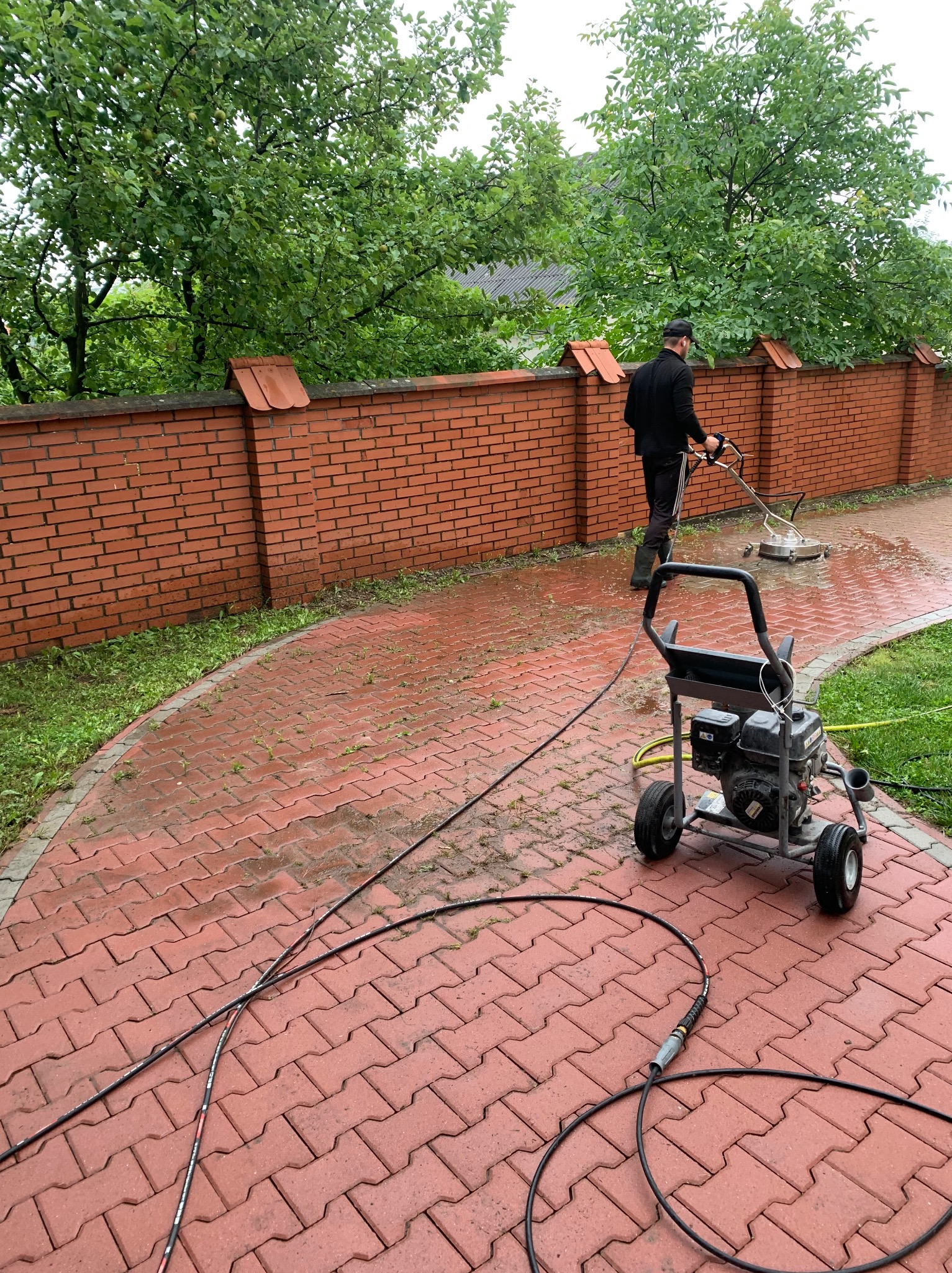 Mężczyzna w czarnym ubraniu czyści czerwoną kostkę brukową na podwórku za pomocą myjki ciśnieniowej z okrągłą dyszą, widoczny mur z czerwonej cegły i drzewa w tle.