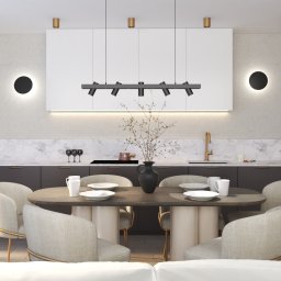 Natura wnętrza Larysa Gesek - Elegancka jadalnia w nowoczesnym stylu z owalnym stołem, beżowymi krzesłami i czarnym wazonem z suchymi gałązkami na tle białych szafek kuchennych, marmurowego blatu i czarnych lamp ściennych.