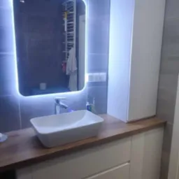 Nowoczesna łazienka z drewnianym blatem, białą umywalką nablatową, podświetlanym lustrem i szafką do sufitu, wykończona szarymi płytkami.