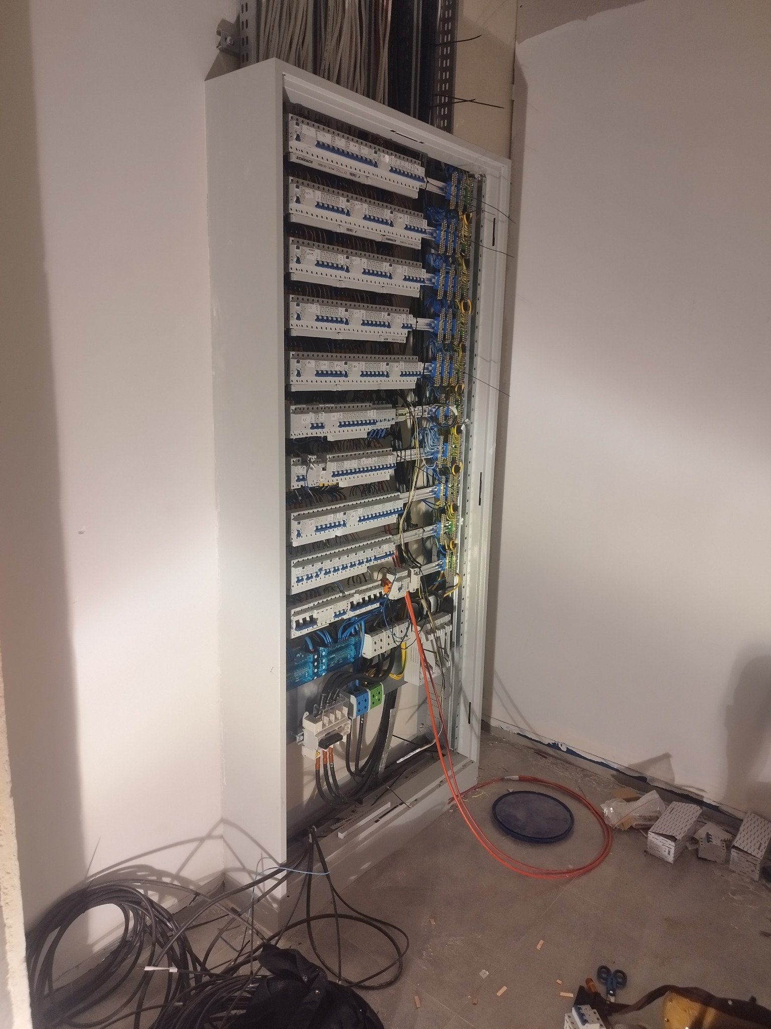 Otwarta szafa elektryczna z licznymi bezpiecznikami i okablowaniem, widoczne kable na podłodze, wnętrze w trakcie montażu.