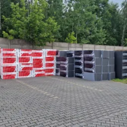 Skladowisko materiałów izolacyjnych: czerwone i szare paczki styropianu EPS Perfect 040 i EPS Grafit 031 ułożone w rzędach na brukowanym placu, z betonowym murem i zielenią w tle.