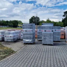 Palety z materiałami budowlanymi, zabezpieczone folią, ustawione na brukowanym placu; w tle widoczna zieleń, błękitne niebo z chmurami i zaparkowana ciężarówka.