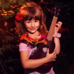 Uśmiechnięta dziewczynka w różowej sukience trzyma ukulele na tle kwitnącego krzewu z pomarańczowymi kwiatami.