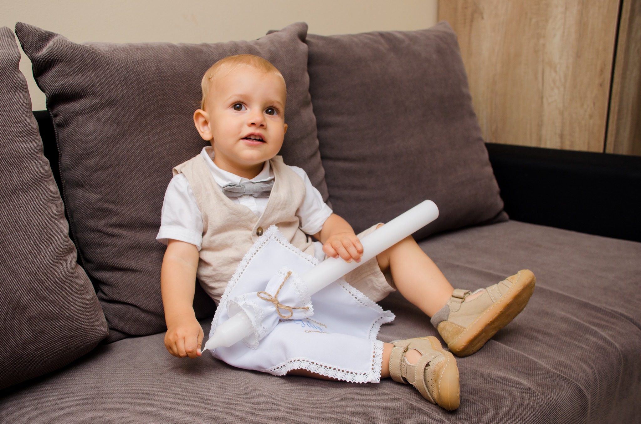 Ubrany w elegancką kamizelkę i muchę chłopiec siedzi na szarej kanapie, trzymając świecę do chrztu owiniętą ozdobną, białą tkaniną z niebieskim napisem.