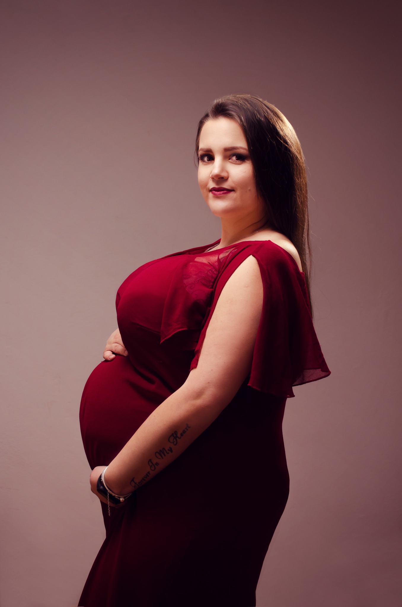 Kobieta w czerwonej sukni ciążowej, trzymająca ręce na brzuchu, z widocznym tatuażem na ramieniu na jednolitym tle.
