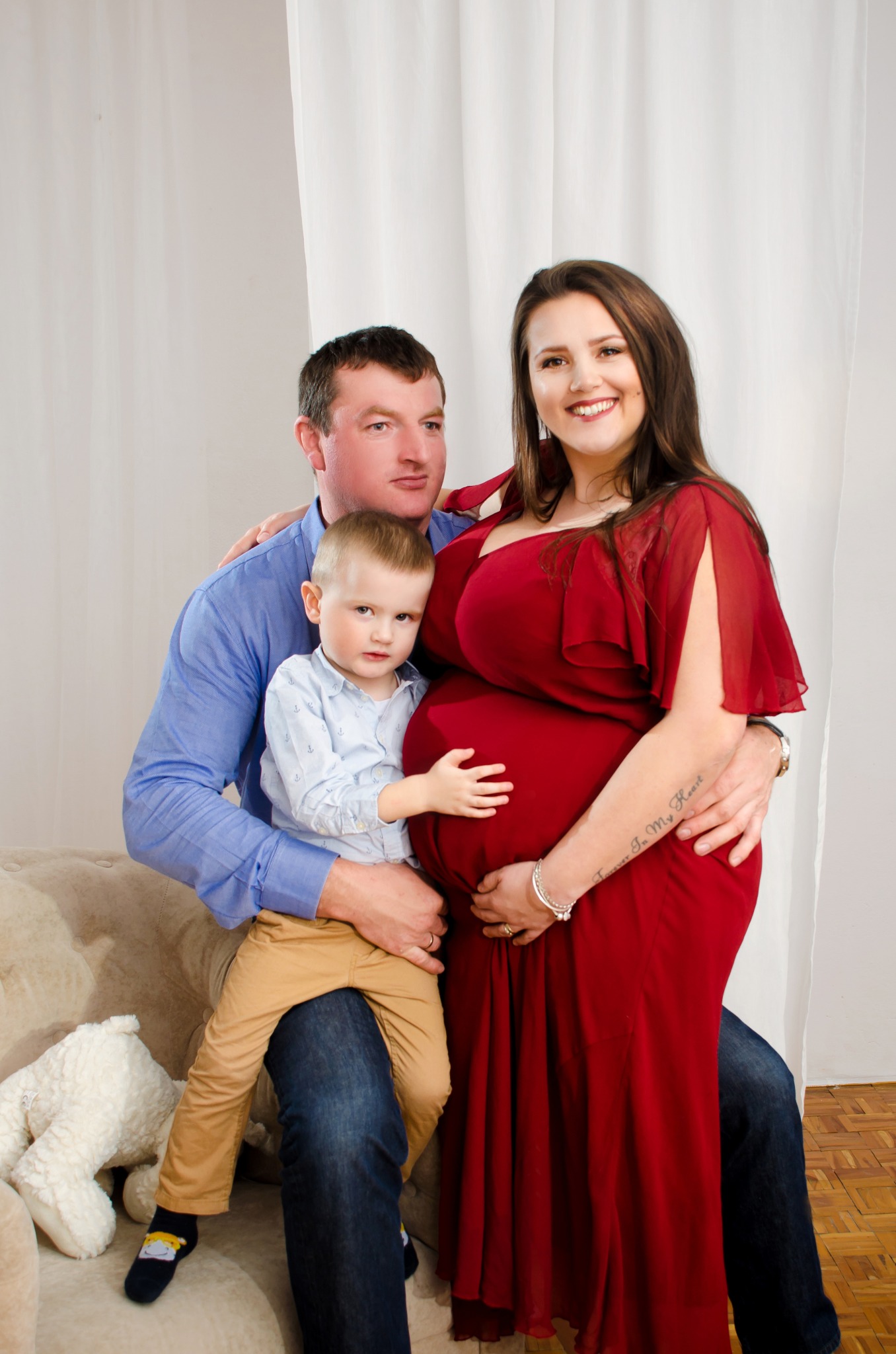 Uśmiechnięta ciężarna kobieta w czerwonej sukience, obejmowana przez męża i synka, który trzyma dłoń na jej brzuchu, na jasnym tle z pluszowym misiem.