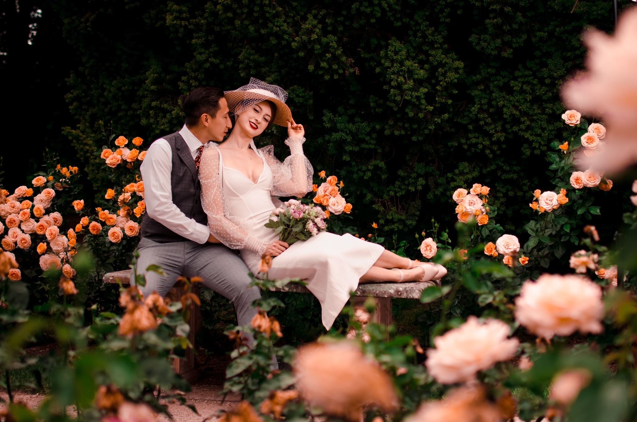 Para w strojach retro siedzi na kamiennej ławce w ogrodzie różanym. Kobieta w białej sukience, kapeluszu i baletkach trzyma bukiet kwiatów. Mężczyzna w kamizelce obejmuje ją ramieniem.