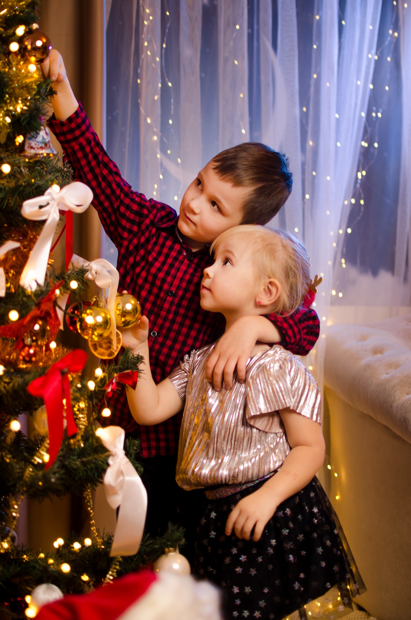 Brat i siostra dekorują choinkę złotymi bombkami i białymi wstążkami, w tle zasłony z lampkami.
