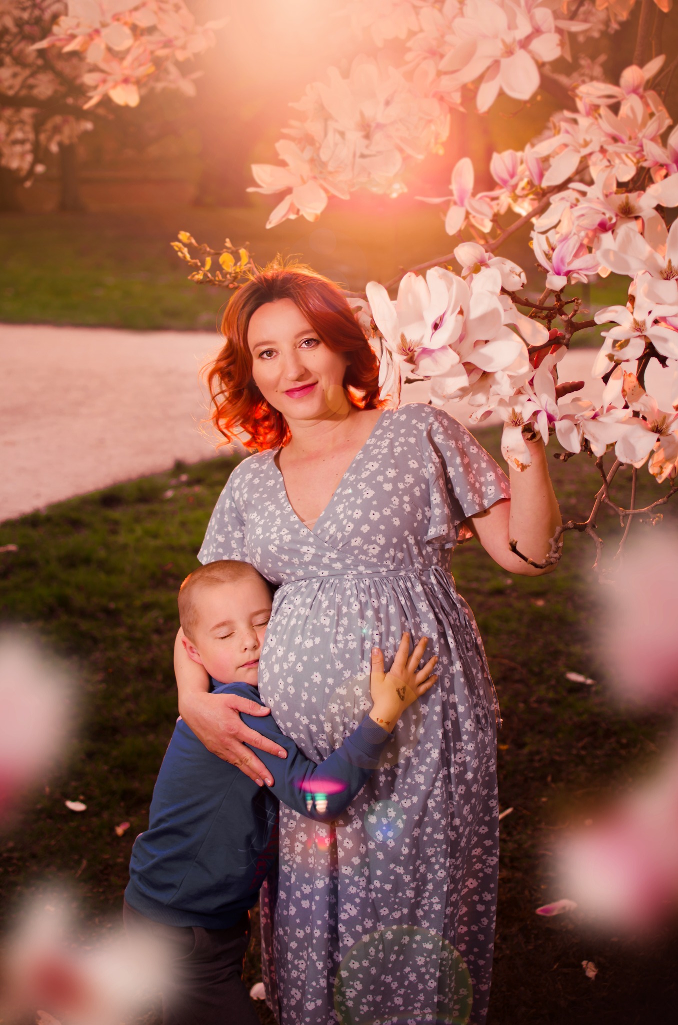 Uśmiechnięta, ruda kobieta w kwiecistej sukience ciążowej, pozuje z synem, który przytula się do jej brzucha na tle kwitnącej magnolii w parku.