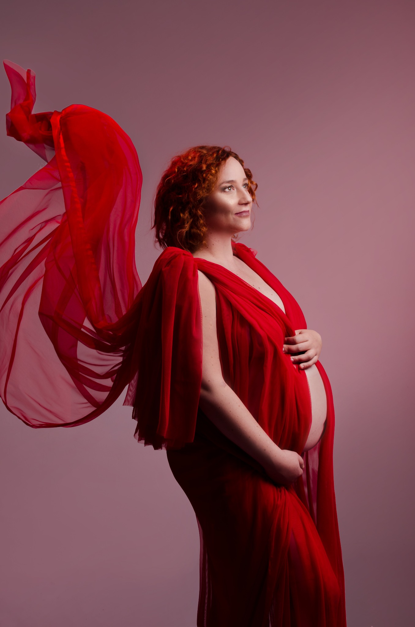 Kobieta w ciąży, otulona zwiewną czerwoną tkaniną, patrzy w górę na tle różowego studia, z delikatnym uśmiechem i dłońmi na brzuchu.