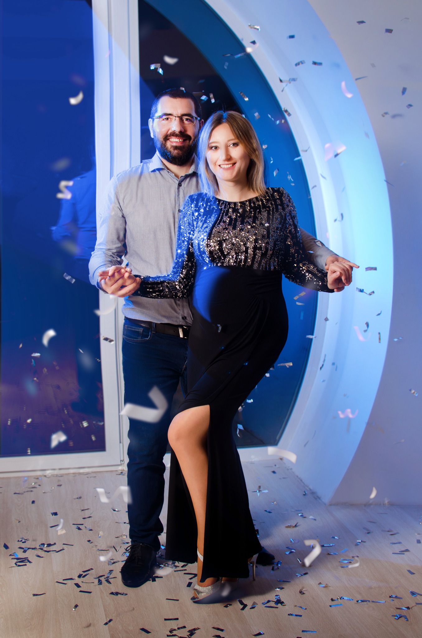 Uśmiechnięta para, kobieta w czarnej sukni z cekinami, mężczyzna w koszuli, otoczeni konfetti na tle okna z niebieskim oświetleniem.