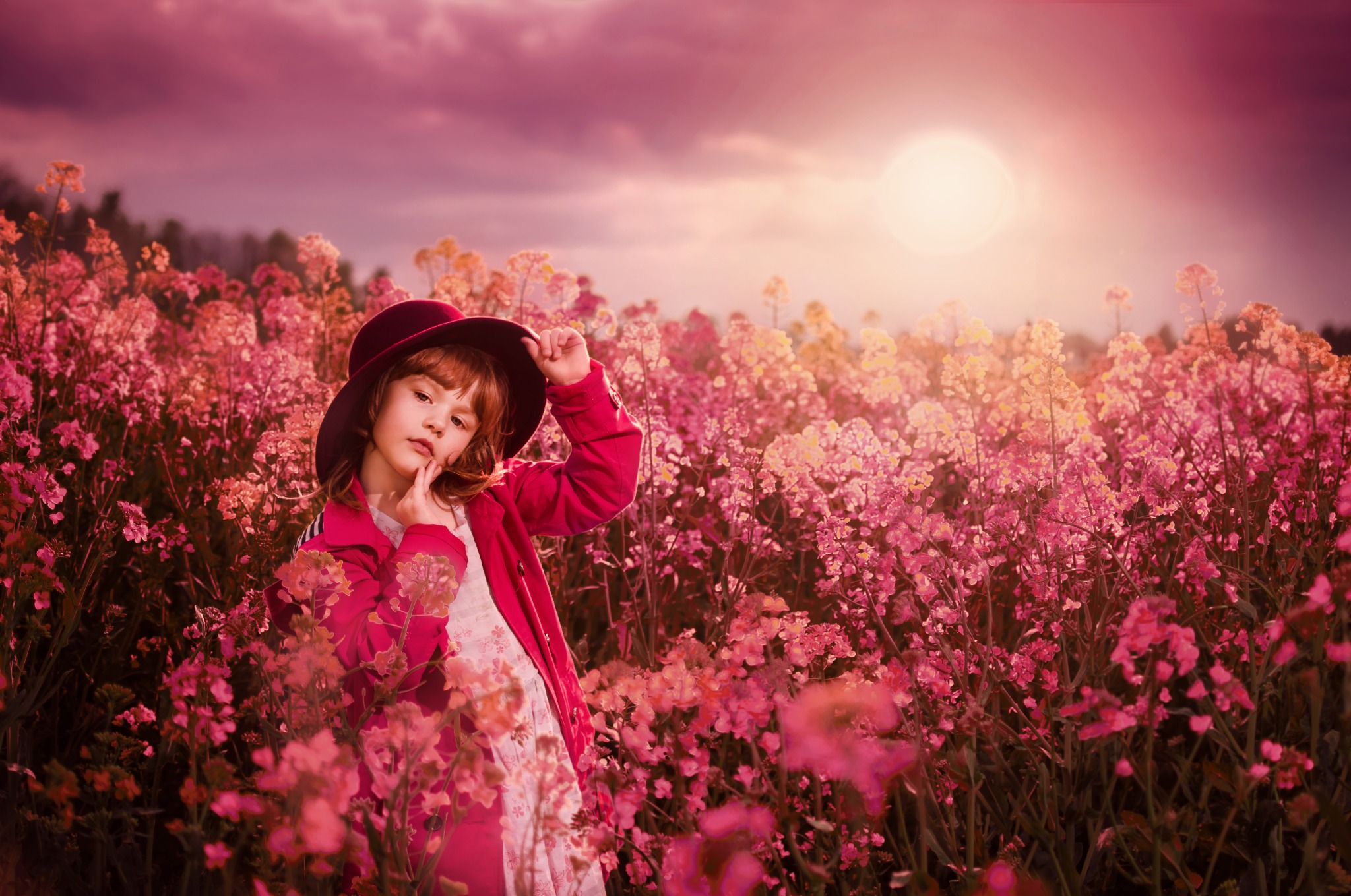 Dziewczynka w bordowym kapeluszu i czerwonej kurtce pozuje w kwitnącym, różowo-żółtym polu, oświetlonym zachodzącym słońcem.