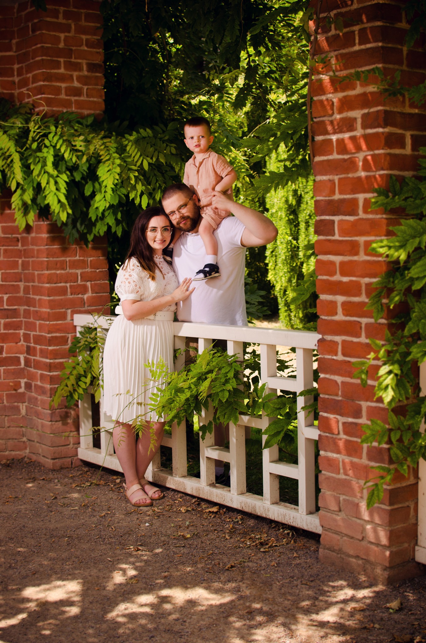 Uśmiechnięta rodzina: kobieta w białej sukience, mężczyzna z synkiem na ramionach, pozują przy ceglanym murze z zielonymi pnączami.