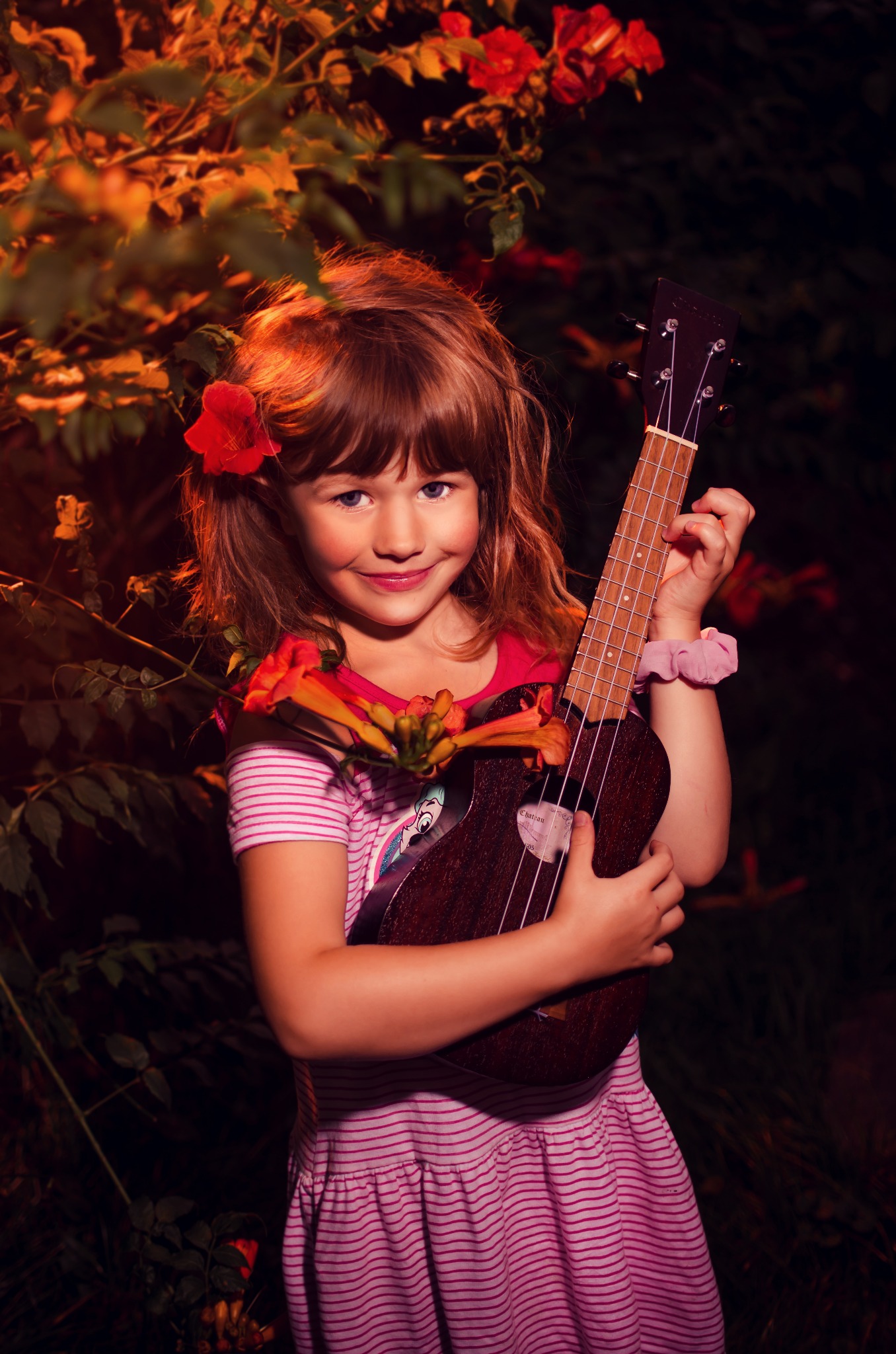 Uśmiechnięta dziewczynka w różowej sukience trzyma ukulele na tle kwitnącego krzewu z pomarańczowymi kwiatami.