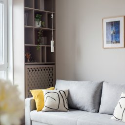 Joanna Tołwińska Fotografka - Jasnoszara sofa z żółtą i wzorzystymi poduszkami, drewniany regał z roślinami, obraz na ścianie i puszysty dywan w minimalistycznym salonie.