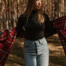 Joanna Tołwińska Fotografka - Portret kobiety w czarnym golfie i dżinsach, na tle leśnych drzew, z zarzuconym na ramiona czerwono-czarnym pledem w kratę, w naturalnym świetle.
