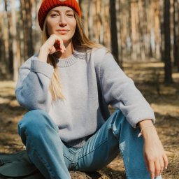 Joanna Tołwińska Fotografka - Portret kobiety w pomarańczowej czapce siedzącej na pniu drzewa w lesie, ubrana w jasny sweter i dżinsy, z plecakiem i butelką obok.