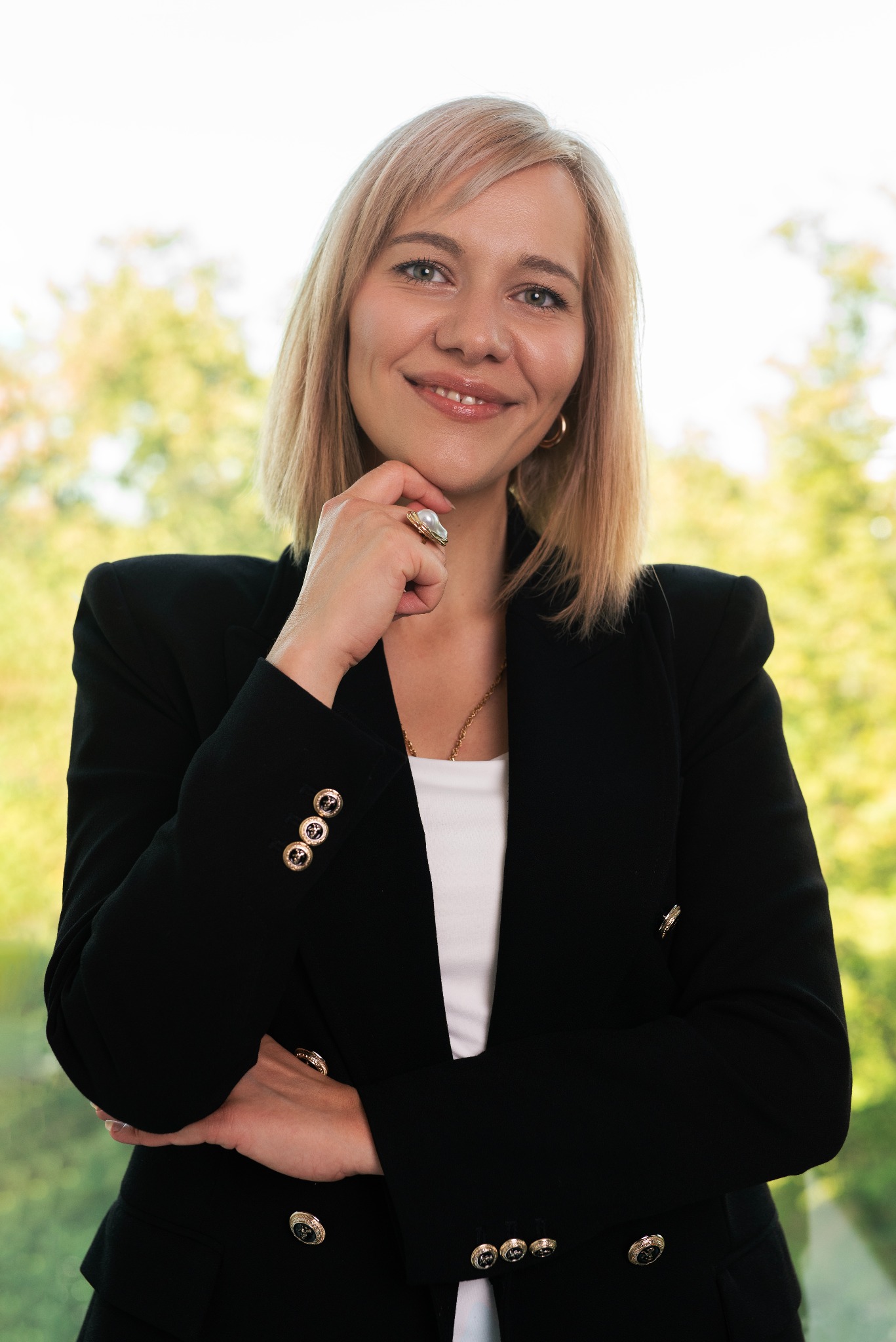 Profesjonalna blond kobieta w czarnej marynarce i białym topie, z dłonią pod brodą i pierścionkiem z perłą, uśmiecha się na tle rozmytej zieleni.