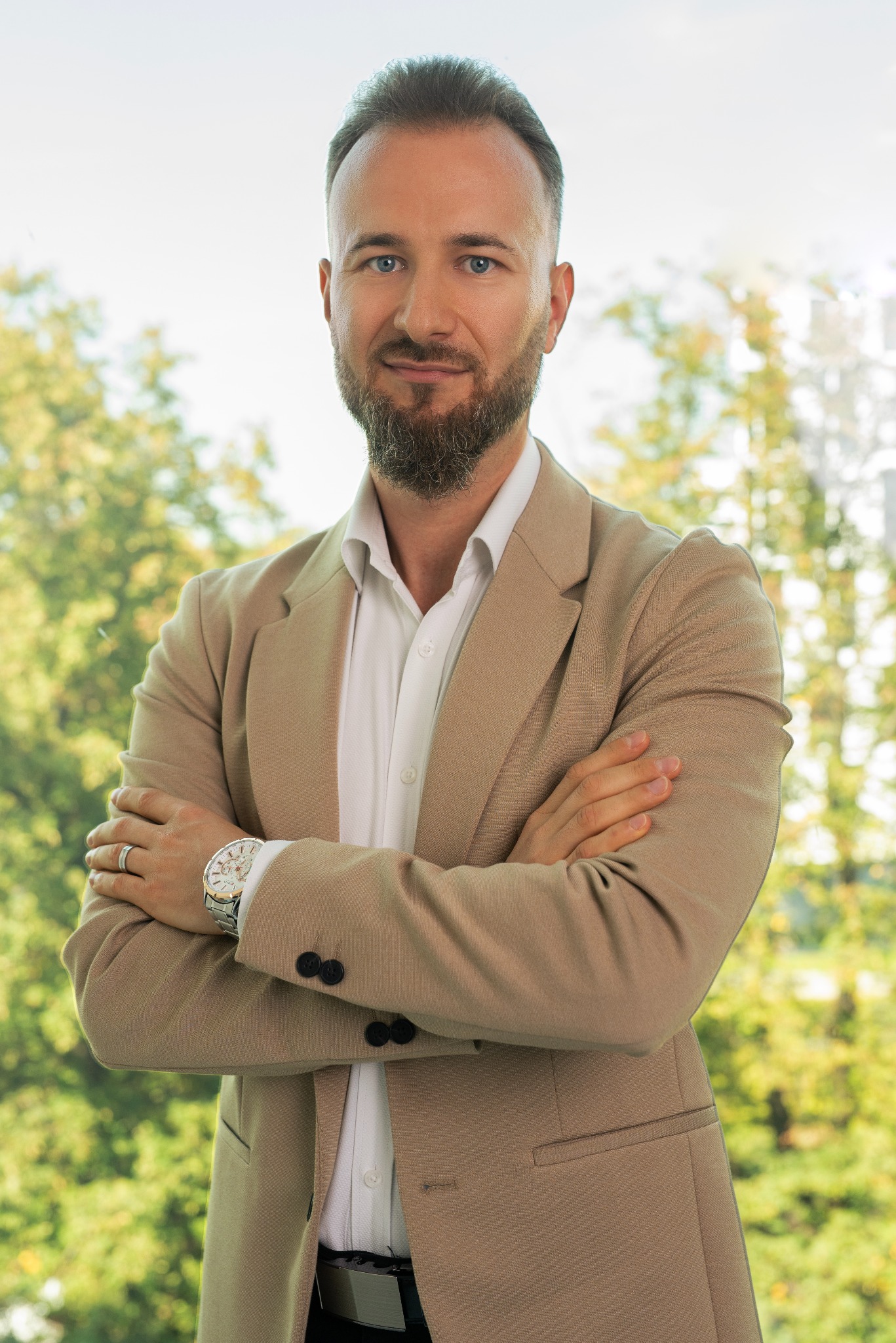 Mężczyzna z brodą w beżowej marynarce i białej koszuli, z zegarkiem na ręku, pozuje na tle zieleni.