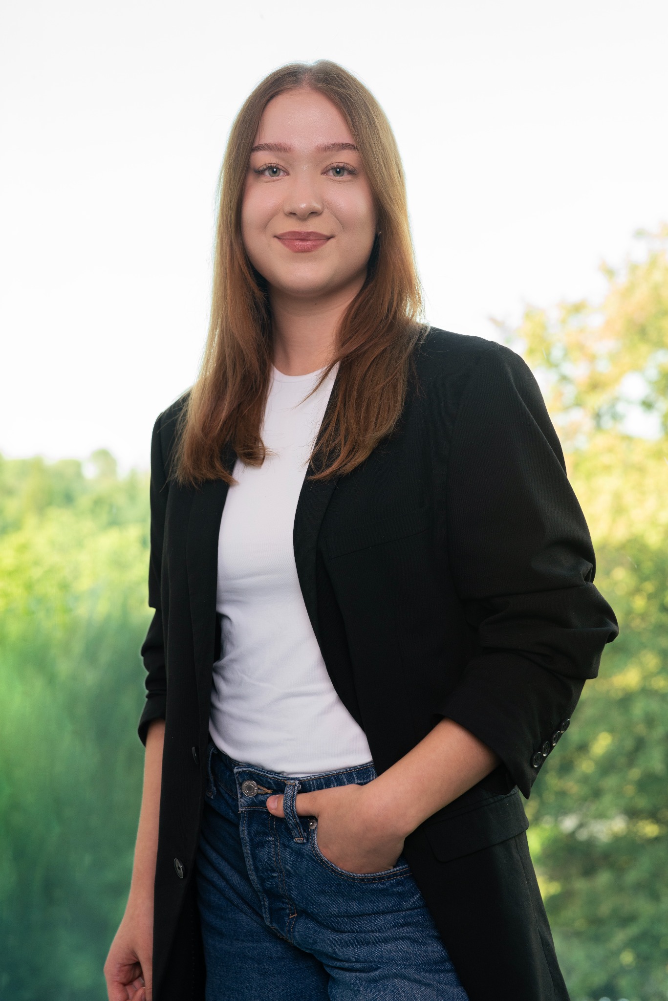 Portret młodej kobiety w czarnej marynarce, białym t-shircie i dżinsach, uśmiechniętej, z ręką w kieszeni, na rozmytym tle zieleni