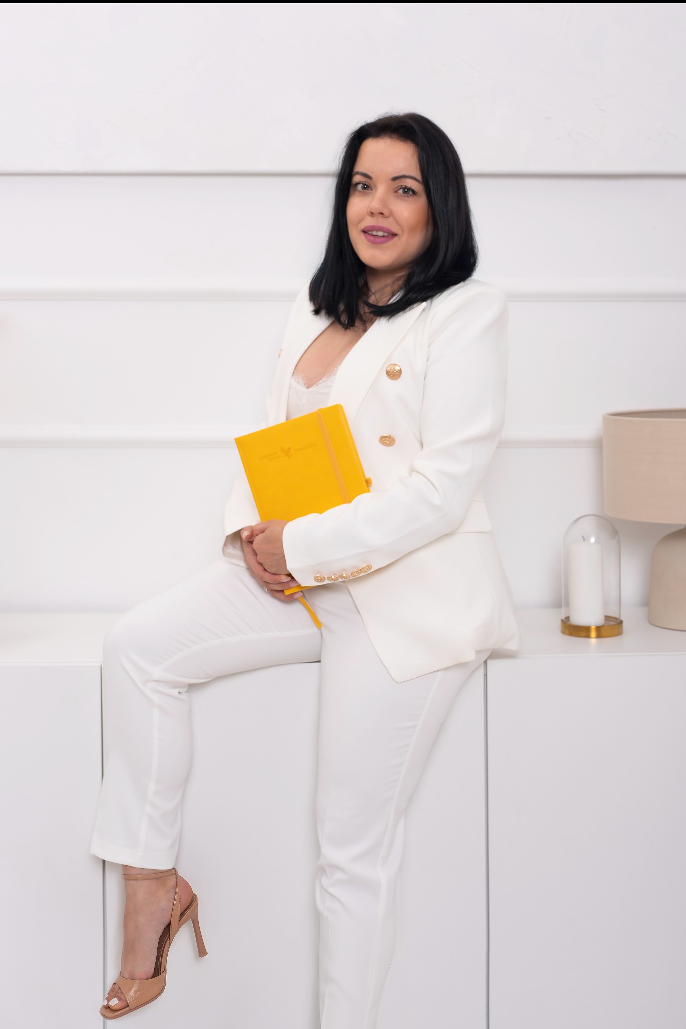 Kobieta w białym garniturze pozuje siedząc, trzymając żółty notatnik z logo, na tle białej ściany z lampą i dekoracyjną świecą.