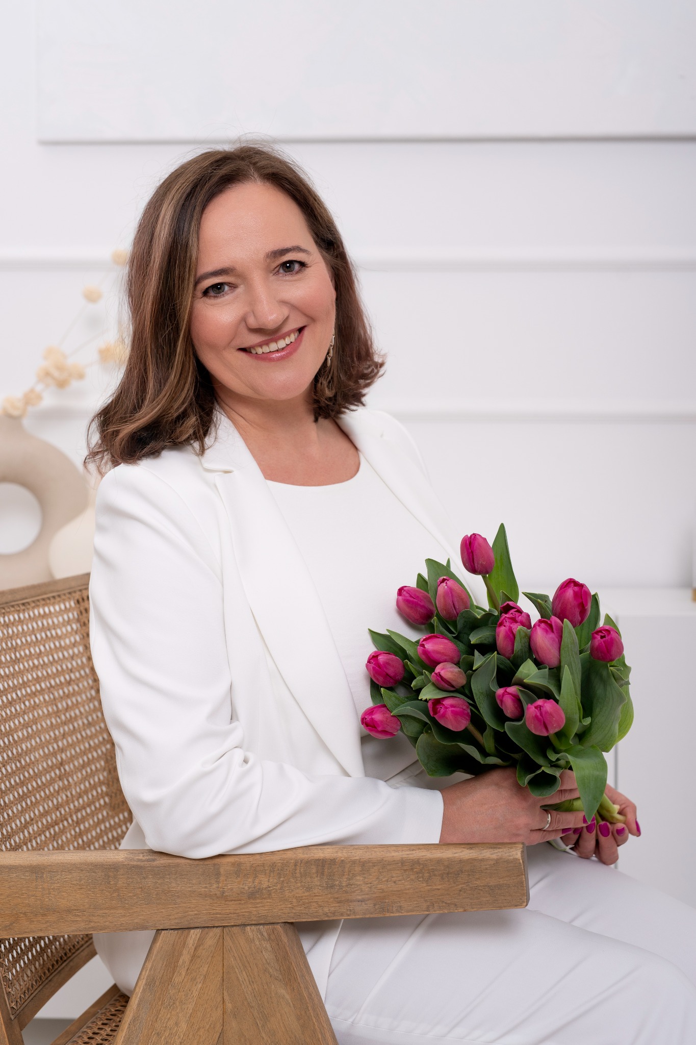 Uśmiechnięta kobieta w białym garniturze siedzi na wiklinowym krześle, trzymając bukiet różowych tulipanów na tle białej ściany z minimalistycznymi dekoracjami.
