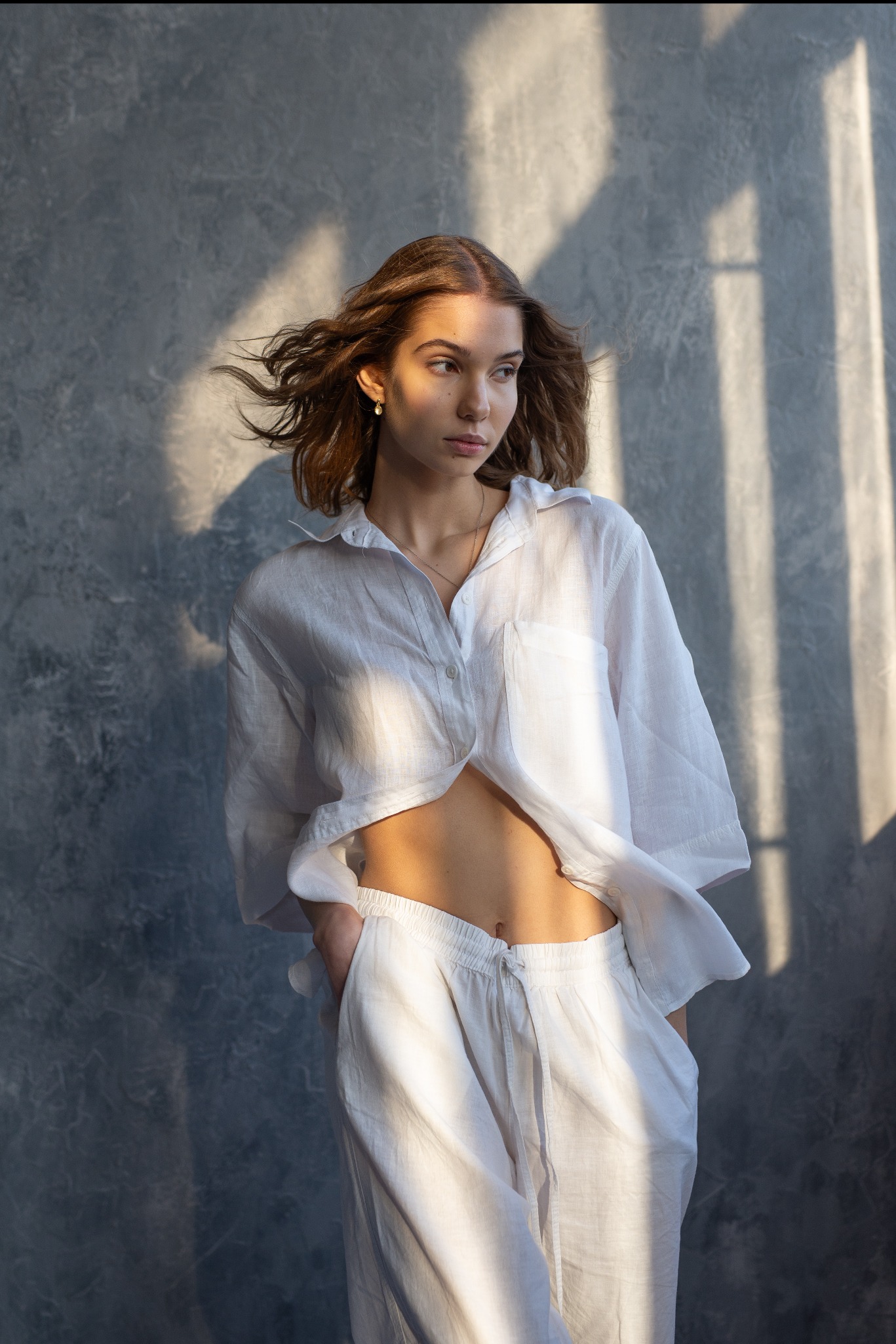 Portret młodej kobiety w białym lnianym komplecie, pozującej na tle szarej ściany z cieniem okiennym, włosy rozwiane, delikatny makijaż.